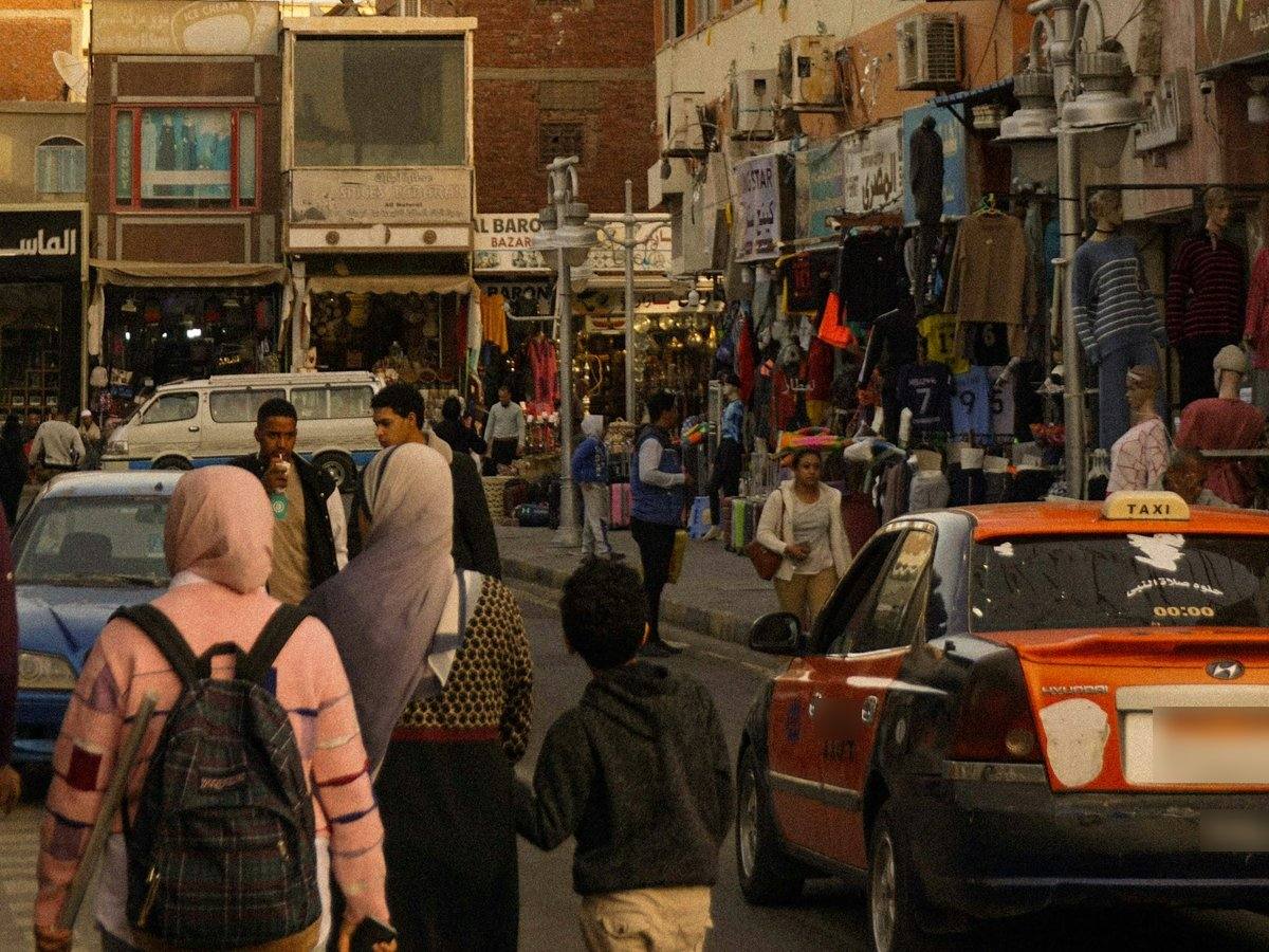 Belebte Einkaufsstraße in Hurghada mit Kleidergeschäften, Passanten und einem orangefarbenen Taxi