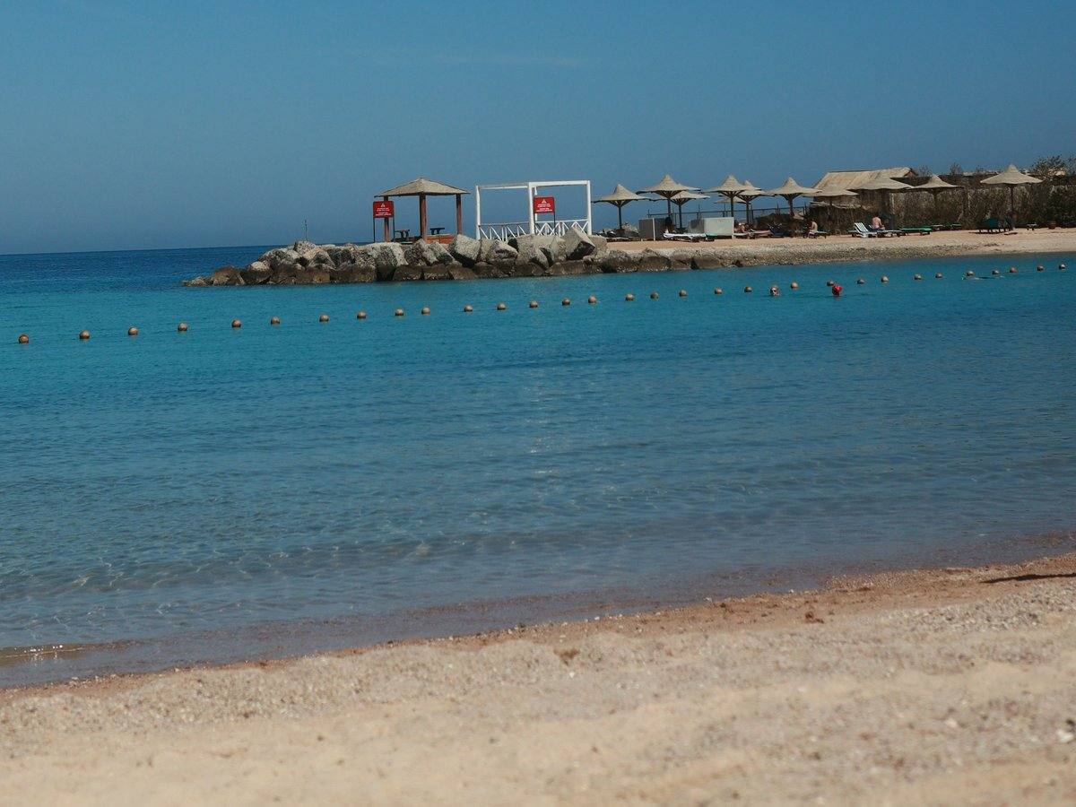 Klares, türkisblaues Wasser an einem Sandstrand in Hurghada, dahinter eine Bojenkette und eine Mole mit Sonnenschirmen
