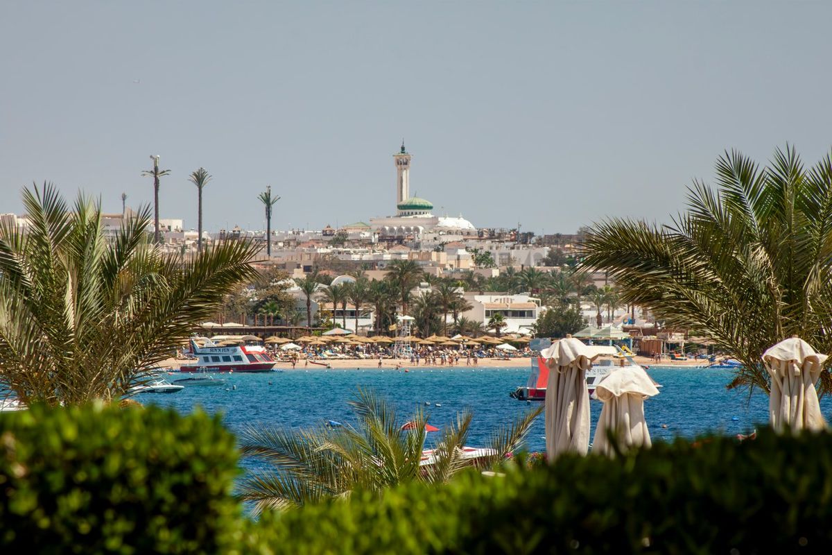 Blick zwischen Palmen auf einen Strand, Boote und die Stadt Hurghada mit Minarett