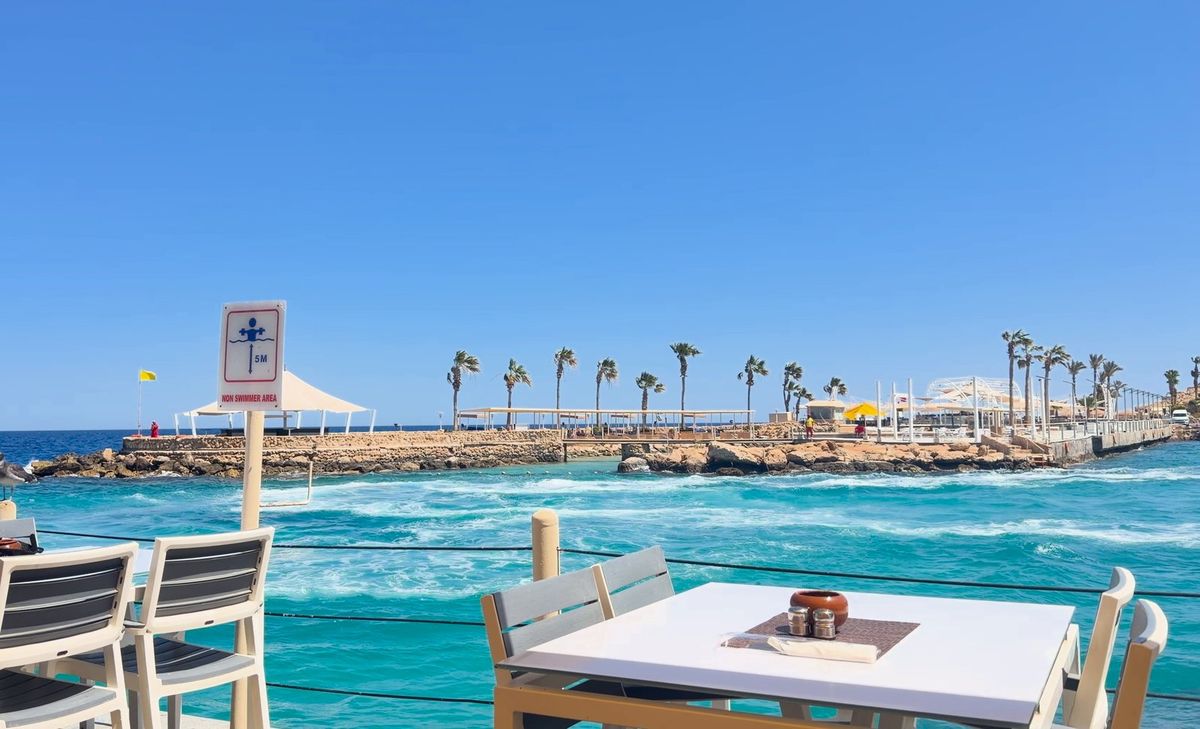 Wellen und Wind am Roten Meer, im Vordergrund eine Restaurantterrasse, hinten ein Steg mit Palmen