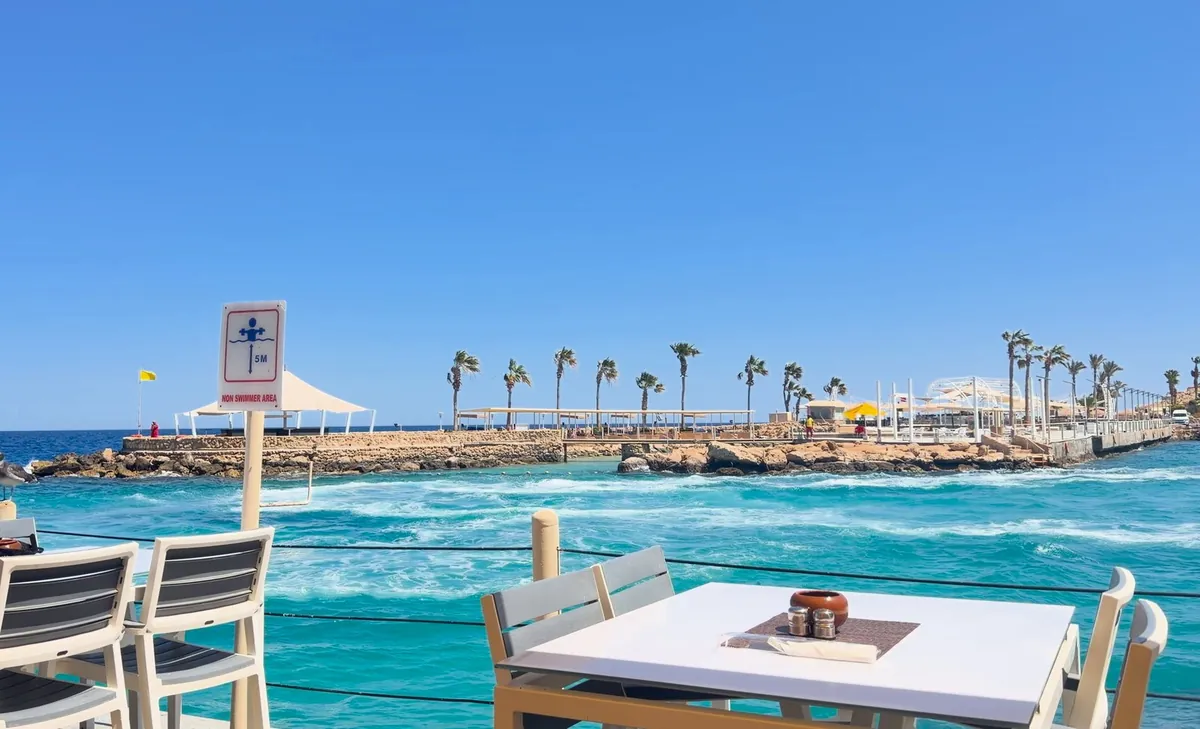Wellen und Wind am Roten Meer, im Vordergrund eine Restaurantterrasse, hinten ein Steg mit Palmen