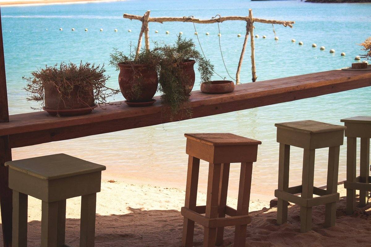Holztheke mit Pflanzentöpfen und Barhockern im Sand einer Strandbar in Hurghada, dahinter türkisfarbenes Meer