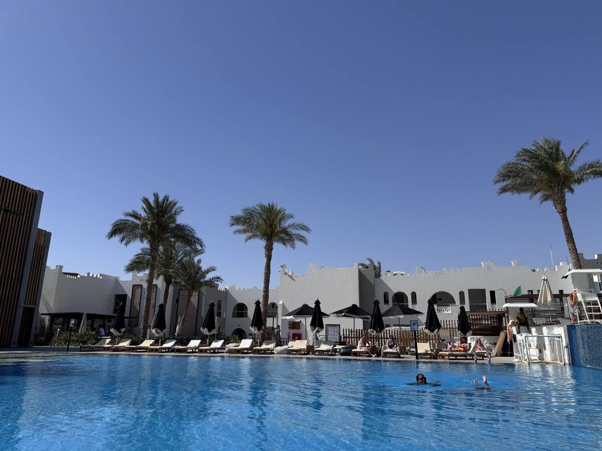 Großer Pool vor weißen Gebäuden mit Palmen und Sonnenschirmen unter blauem Himmel