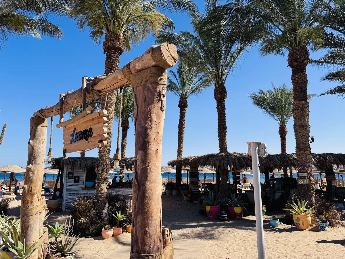 Holzschild Lounge am Eingang einer Strandbar mit Palmen und Sonnenschirmen am Meer